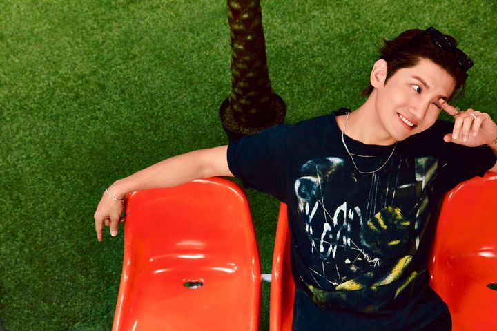 黒いTシャツ姿のチャンミンが赤い椅子に腰掛け、笑顔で目元に手を添えている