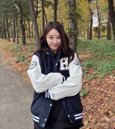 漢陽大学の学生ジャンパーを着て、木々の並ぶ屋外で笑顔を見せるチョン・ユミ。
