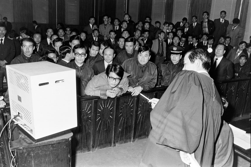 1974년 11월 13일 서울고등법원 법정에서 정치근(오른쪽 법복 입은 이) 부장검사가 육영수 여사 피습 상황을 담은 영상을 설명하고 있다.수갑을 찬 피고인 문세광(가운데)이 영상을 보며 당황한 표정을 짓고 있다.