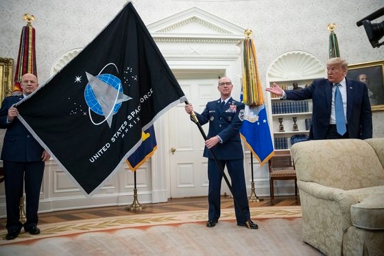 2020년 5월 15일 미국 워싱턴 DC 백악관 집무실에서 열린 우주군기 수여식에서 당시 도널드 트럼프(오른쪽) 대통령의 제스처에 맞춰 존 레이먼드(왼쪽) 우주군참모총과 로저 토버먼 주임원사가 우주군 깃발을 들어보이