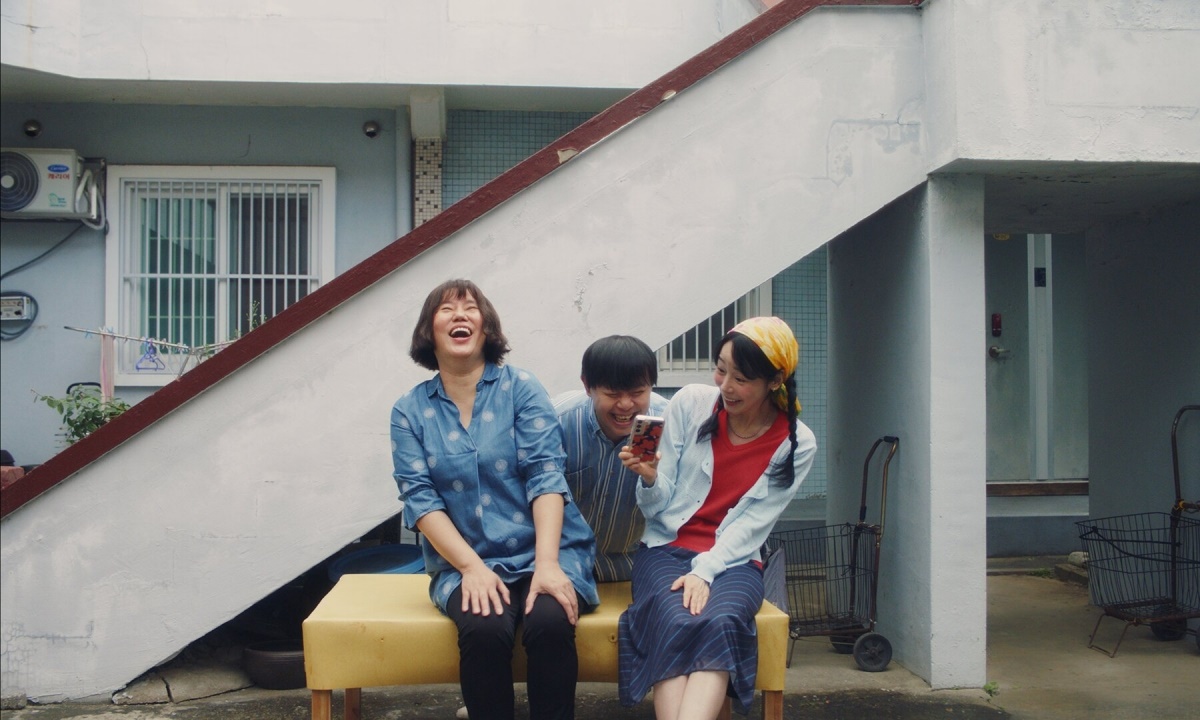 映画『フルムーン』で、集合住宅の前に座って笑い合う母娘と男性の姿