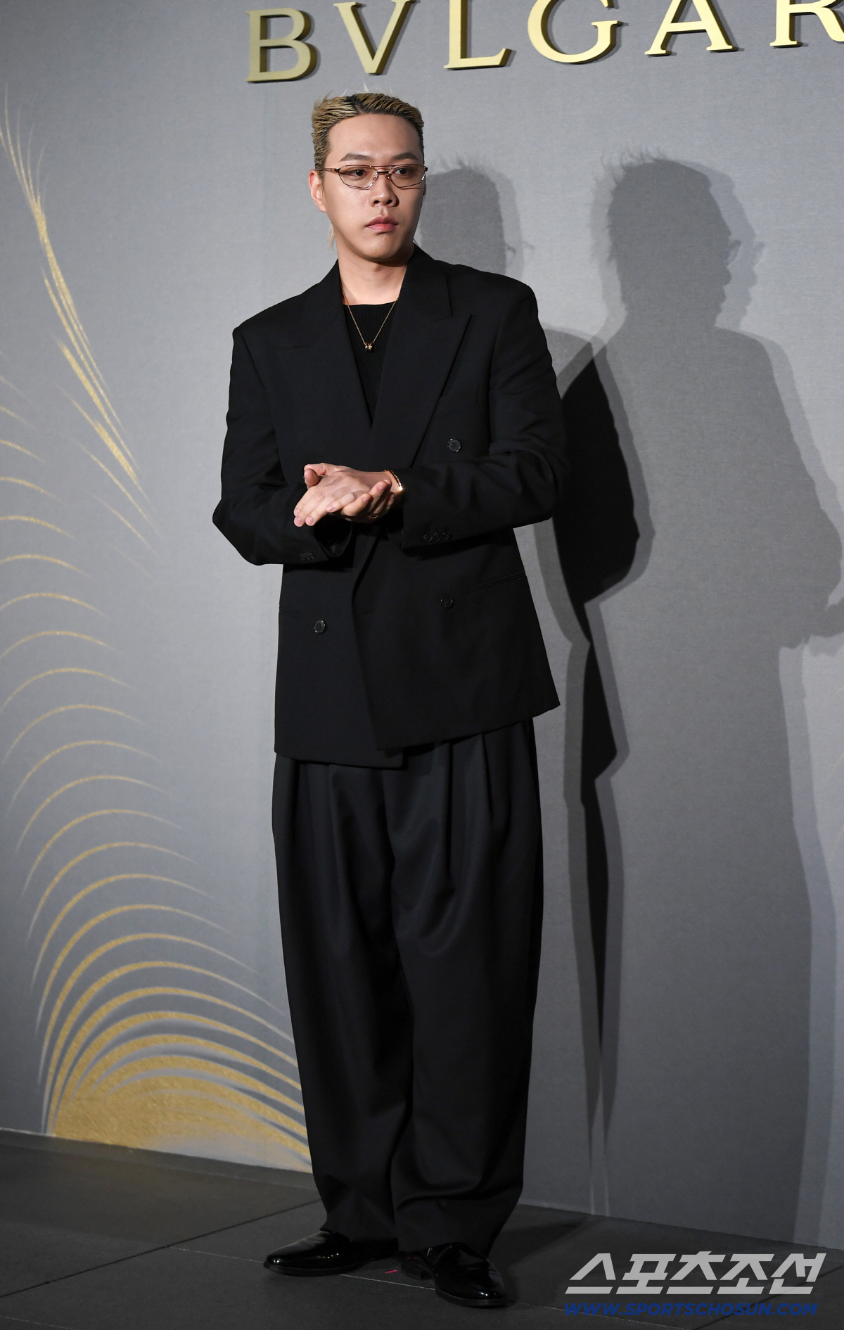 ブルガリのロゴを背に、黒いスーツと眼鏡姿で両手を重ねるBewhY