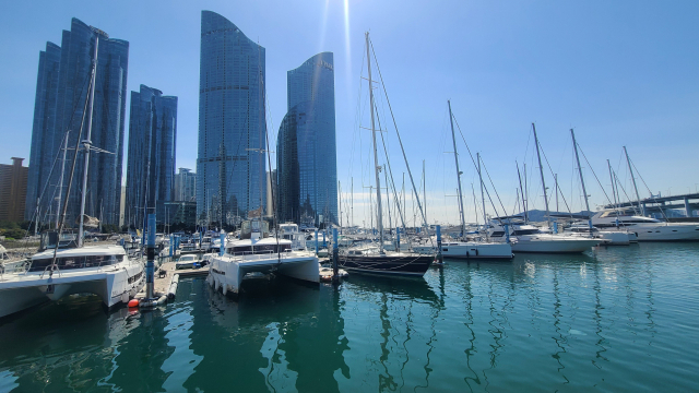 수영만 요트경기장 내 요트 계류장 쪽으로 다가서면, 잔잔한 물결 위에 계류 중인 하얀색 요트들이 바다를 한가득 메우고 있다. 마린시티 초고층 건물의 커튼월 유리창은 하늘을 배경으로 푸른빛을 발하고 있다.
