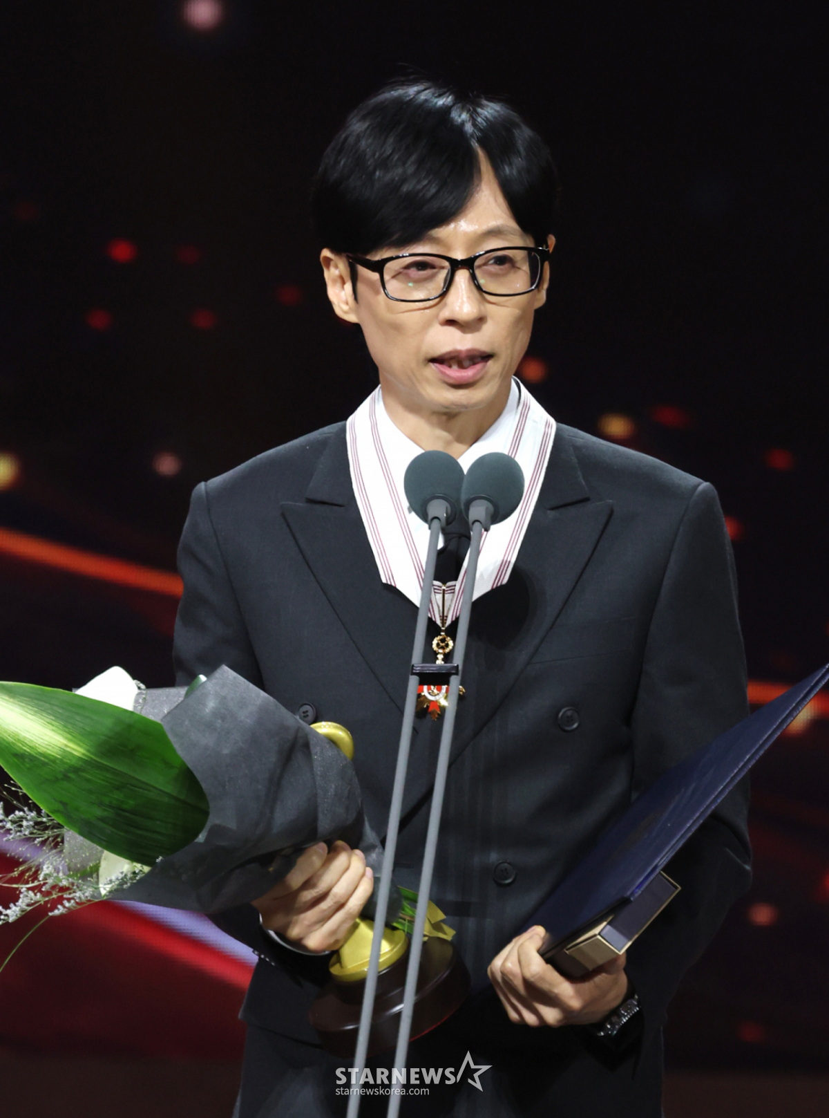 受賞スピーチをするユ・ジェソク。胸元に保管文化勲章を着け、花束と賞状を手にしている