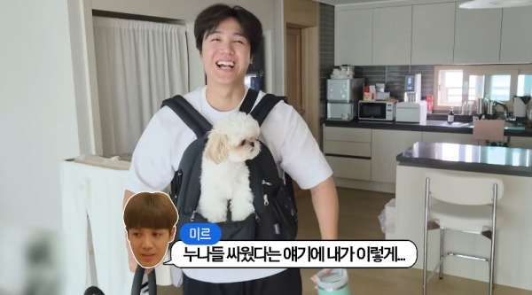 家族チャンネル「방가네」の動画キャプチャ。白い犬を抱っこひもに入れて笑うミルが台所に立ち、画面下に「姉たちがけんかしたって聞いて、僕がこんなふうに…」という字幕が表示されている