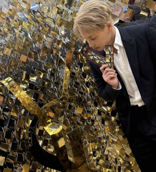 金髪のカイが黒いスーツ姿で、鏡のような金色の壁の前に立つ