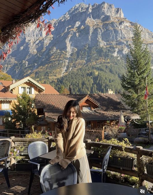 スイスの山々を背景に、屋外のテラスで椅子に手を添えるイ・ソリ