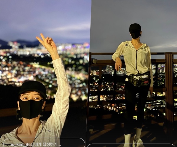 黒いキャップとマスク、白いウインドブレーカー姿で、夜景を背にポーズを取るキム・ヘス