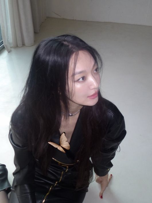 長い黒髪のシン・ミナが、蝶の飾りが付いた黒いレザー風のトップスとチョーカーを着け、床に手をついて横を見ている。