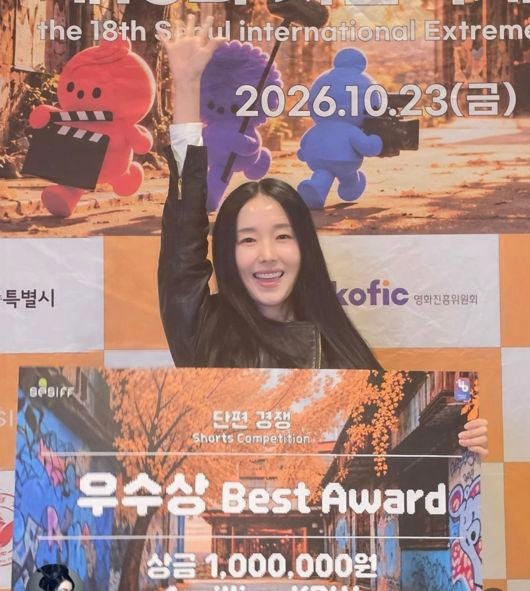 短編映画祭の会場で、優秀賞の賞状を掲げて笑顔を見せるイ・ジョンヒョン。背景には映画祭のパネルが写っている。