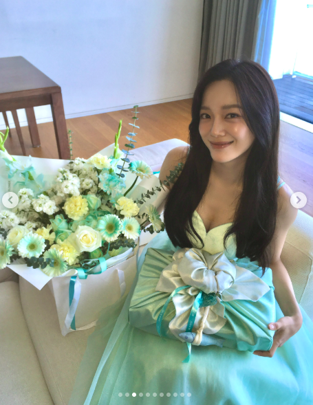 淡いミント色のドレスを着たキム・セジョンが床に座り、花束と包みを抱えて微笑む写真。