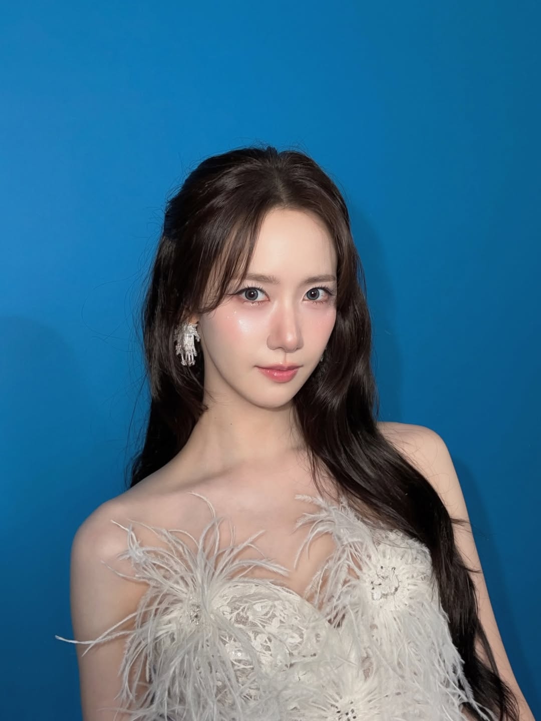 白い羽根をあしらったドレスを着て、青い背景の前でカメラを見つめるイム・ユナ