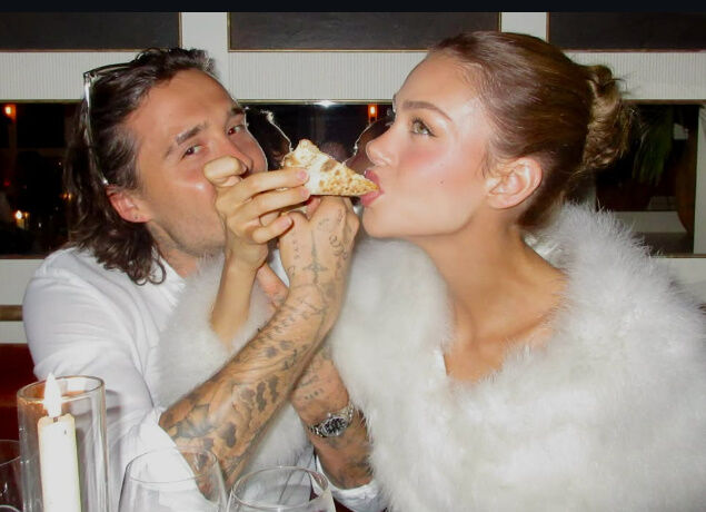 ブルックリン・ベッカムが持つピザに、妻ニコラ・ペルツが顔を寄せている。二人は笑顔で食事を楽しんでいる。