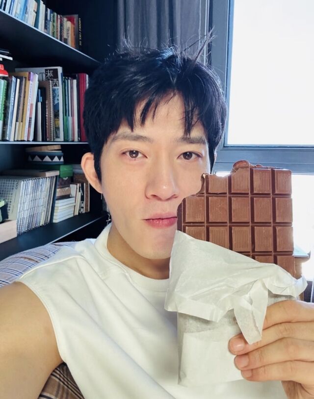 白いノースリーブ姿の井柏然が、板状の大きなチョコレートを顔の横に掲げた自撮り