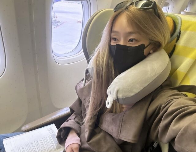 飛行機の窓際の座席で、白いネックピローと黒いマスクを着けたヒョリン