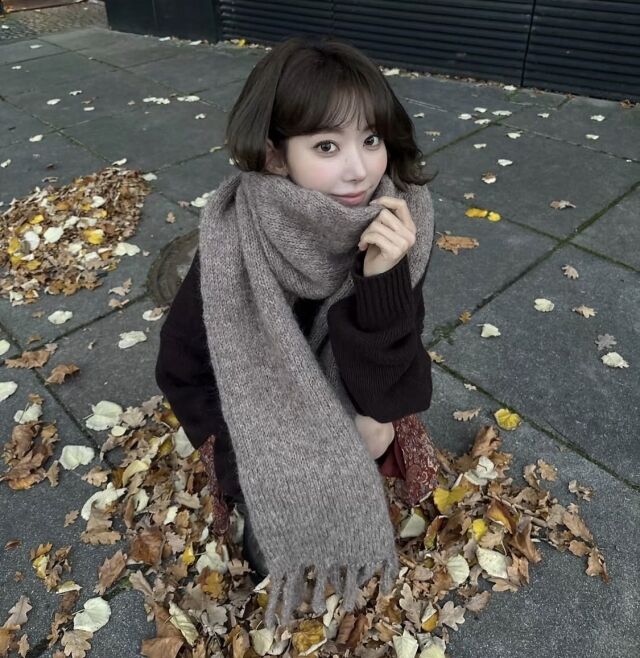 落ち葉のある屋外で、SAKURAが自身で編んだ茶色のマフラーを巻いて座っている