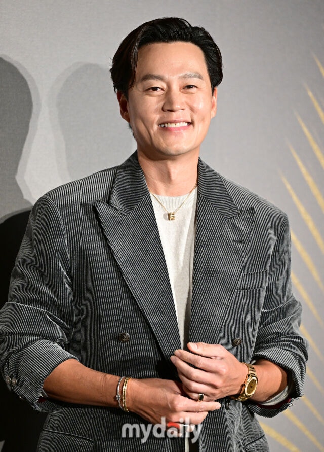 細いストライプ柄のジャケットと白いトップスを合わせたイ・ソジンが、手を重ねて笑顔を見せる。腕時計とブレスレットも着用。