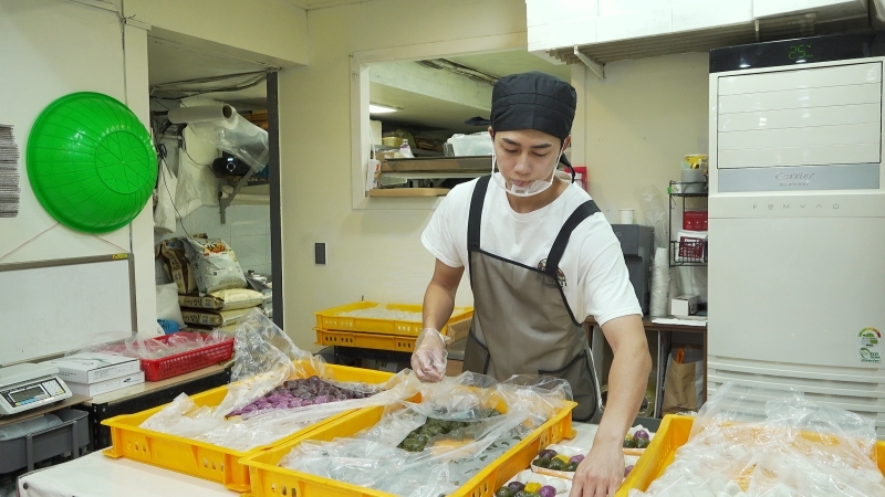 餅店で作業するアベ・シンさん。黄色いトレーに並ぶ餅を整えている。
