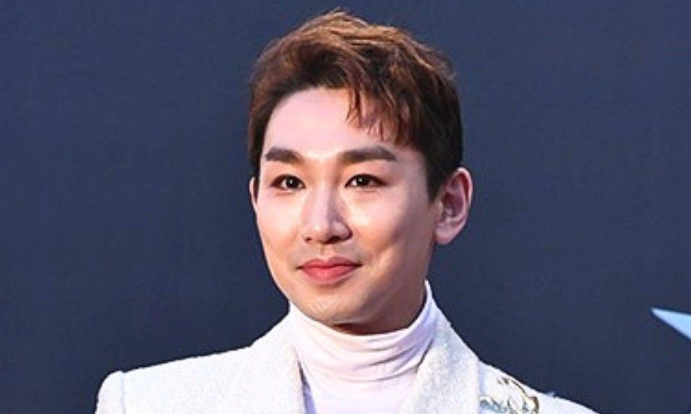 白いタートルネックと白いジャケット姿のキム・ホヨンが、青い背景の前で正面を向く上半身写真