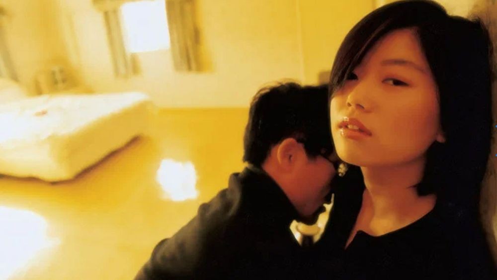 映画『嘘』の一場面。女性と、その背後に座る男性が写っている