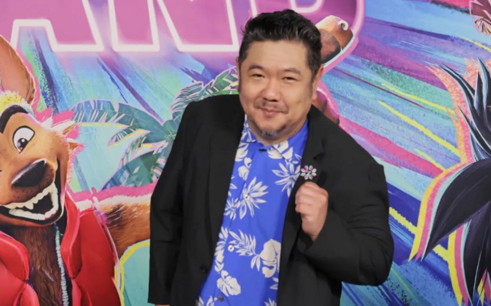 映画の告知パネルを背に、青い花柄シャツと黒いジャケット姿でポーズを取るエリック・バウザ