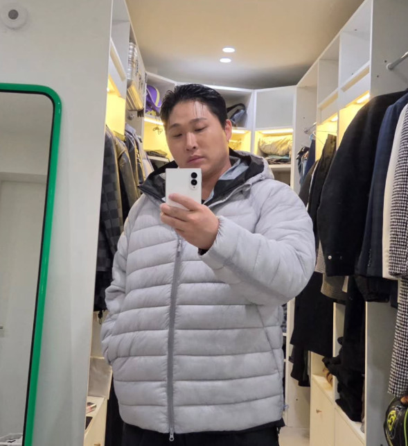 衣類が並ぶ部屋で、グレーのパディングを着て鏡越しに自撮りするスウィングス