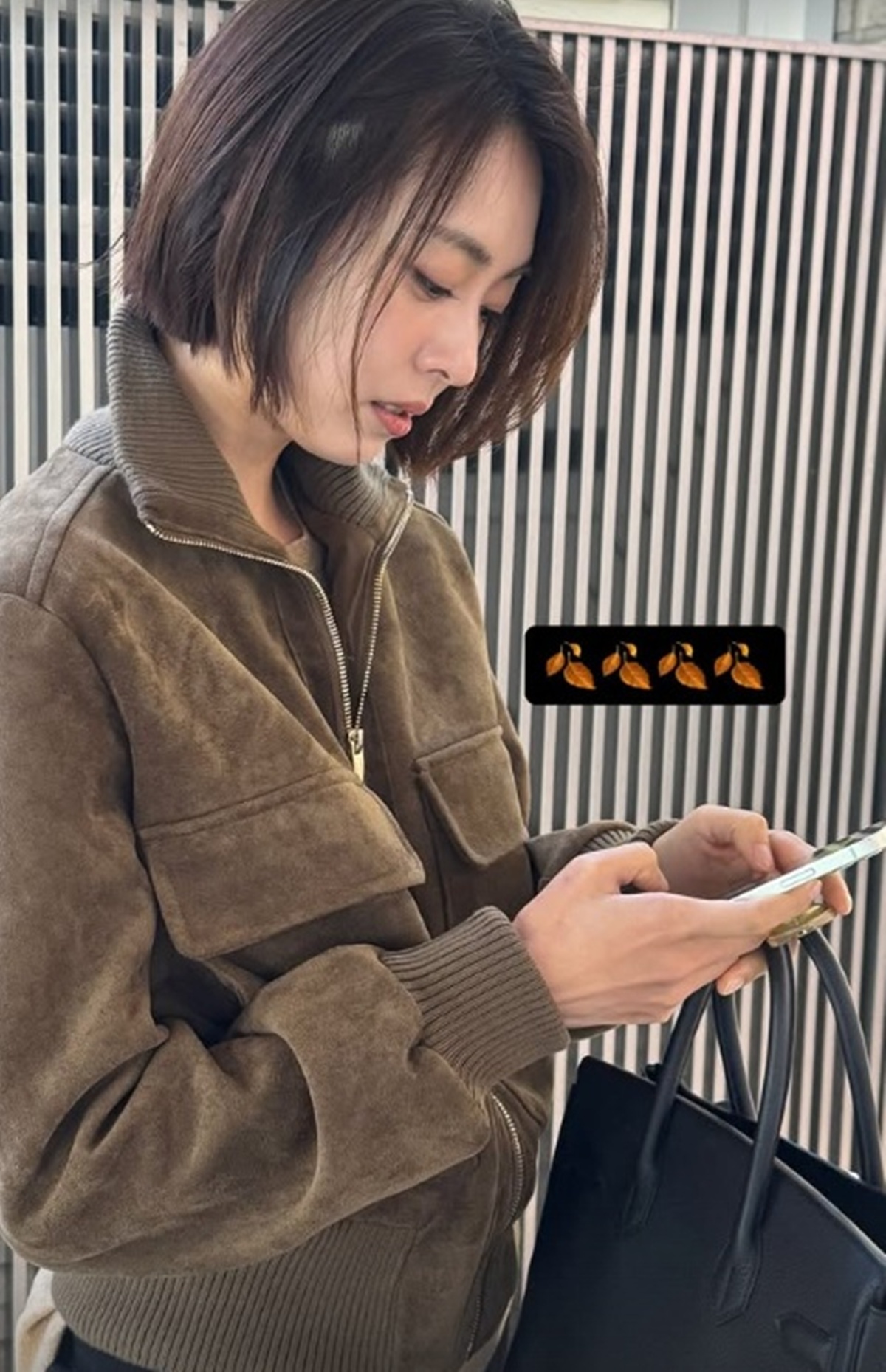 あごのあたりまでのボブヘアで、ブラウンのスエードジャケットを着てスマートフォンを見るイ・ヨニ