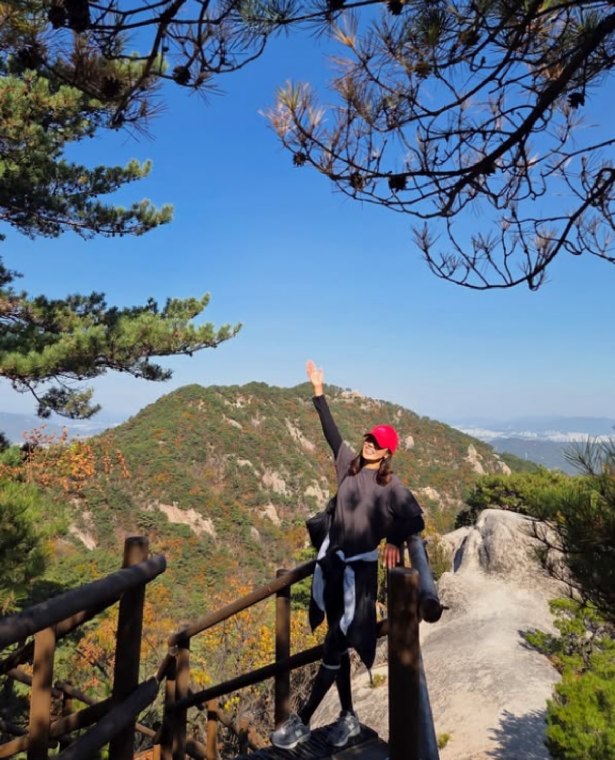 赤い帽子をかぶったチャン・シニョンが、秋色の山々を背景に山頂で片手を上げている