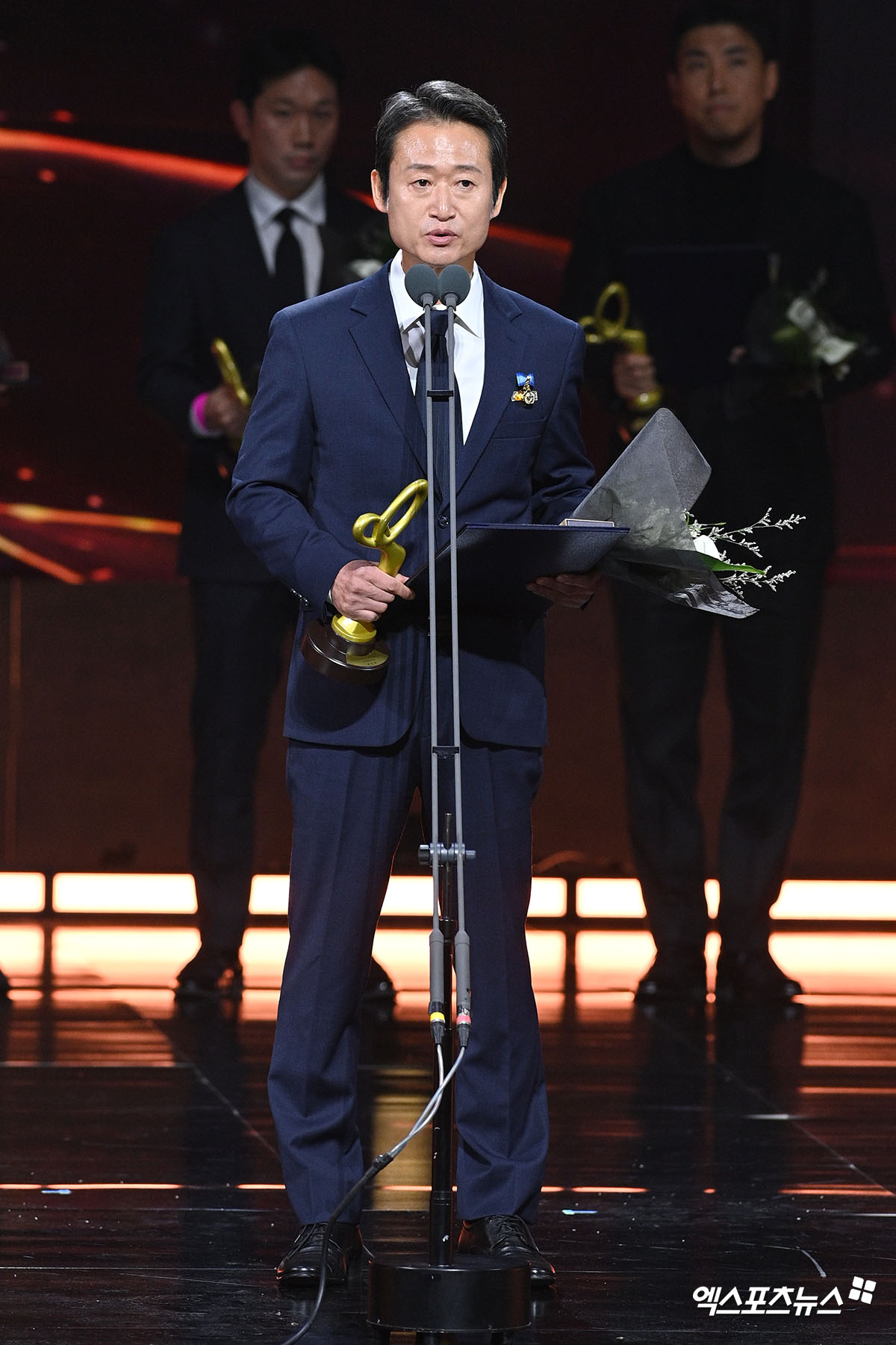 濃紺のスーツ姿で賞牌と花束を持ち、マイクの前で受賞所感を述べる俳優ユ・スンモク