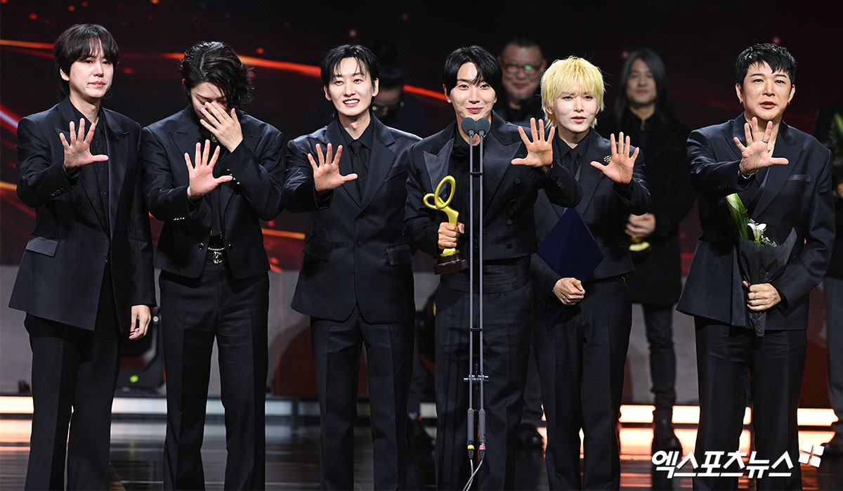 SUPER JUNIORのメンバー6人が舞台で大統領表彰のトロフィーを掲げ、中央のマイク前で受賞後の感想を伝えている