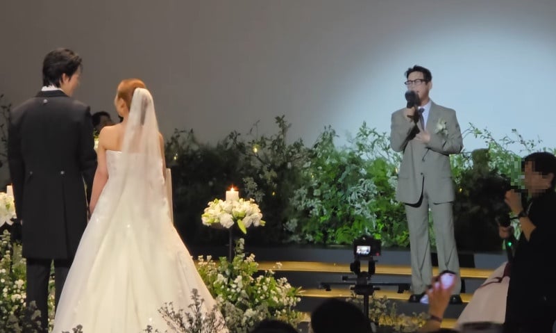 新郎新婦を前に、灰色のスーツ姿の歌手テイがマイクを持って祝歌を歌う結婚式の様子
