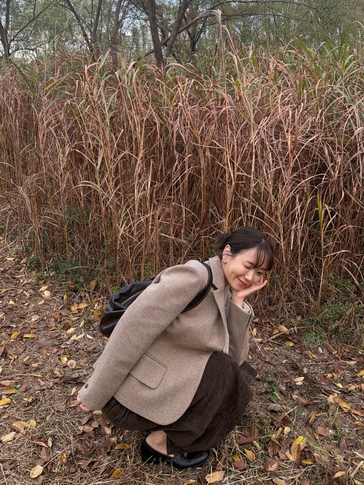 ベージュのジャケットと濃い茶色のロングスカート姿で、ススキを背景に頬へ手を添えて笑う30期オクスン