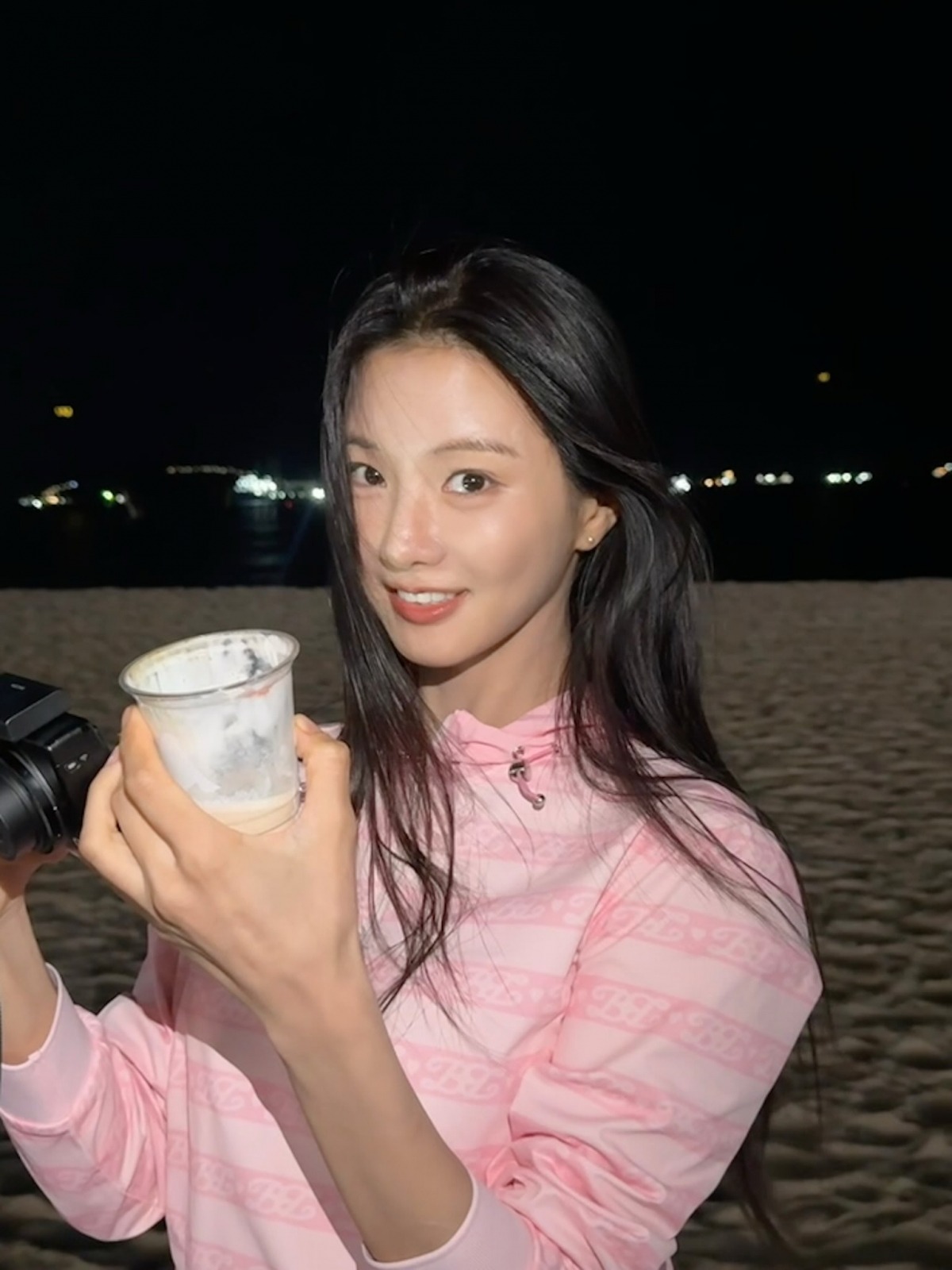 ピンクのフーディー姿のイ・セヒが釜山の夜の海辺でカップを手に笑顔を見せる