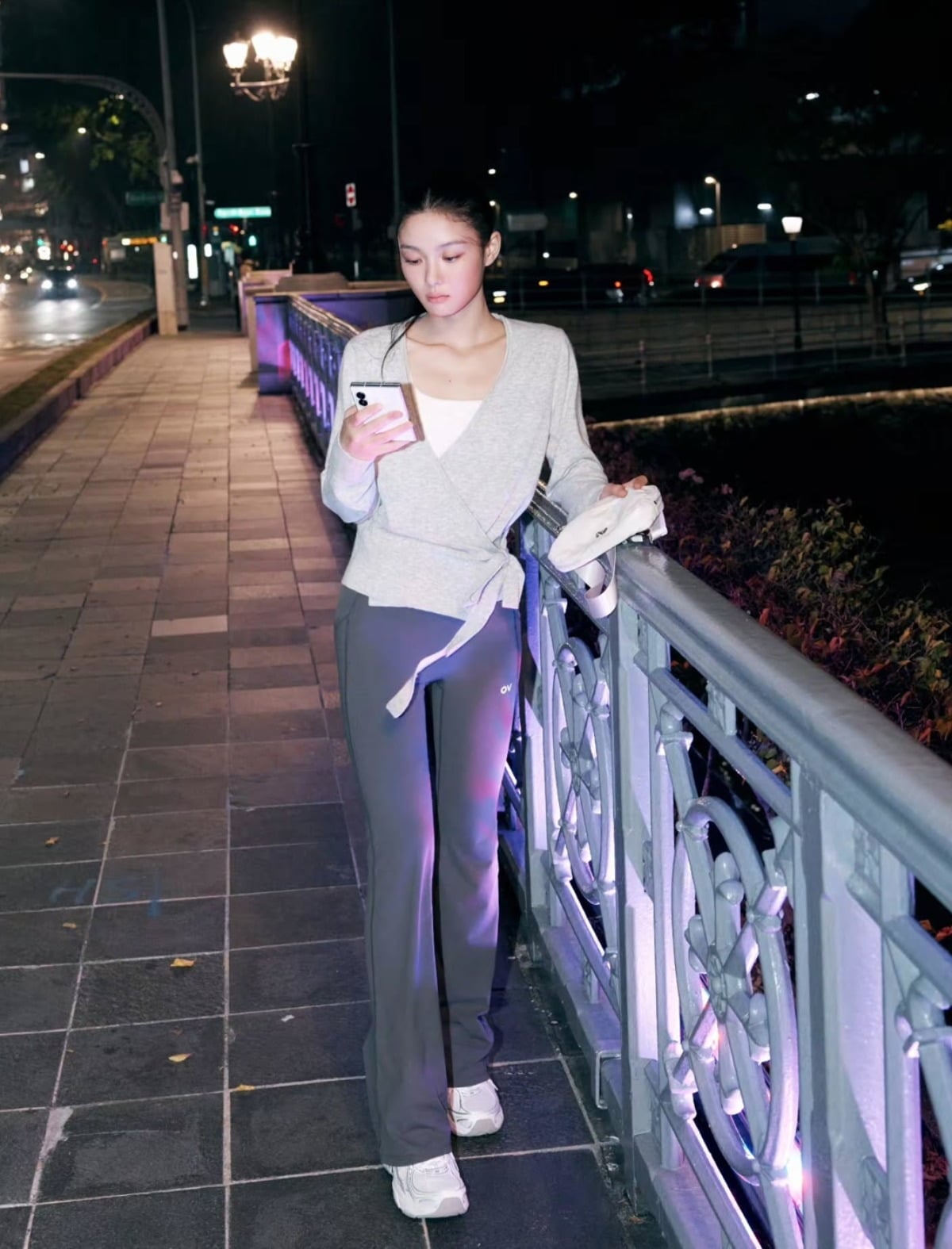 夜の街の手すりに寄りかかり、スマートフォンを見るキム・ユジョン。グレーのトップスとレギンス姿。