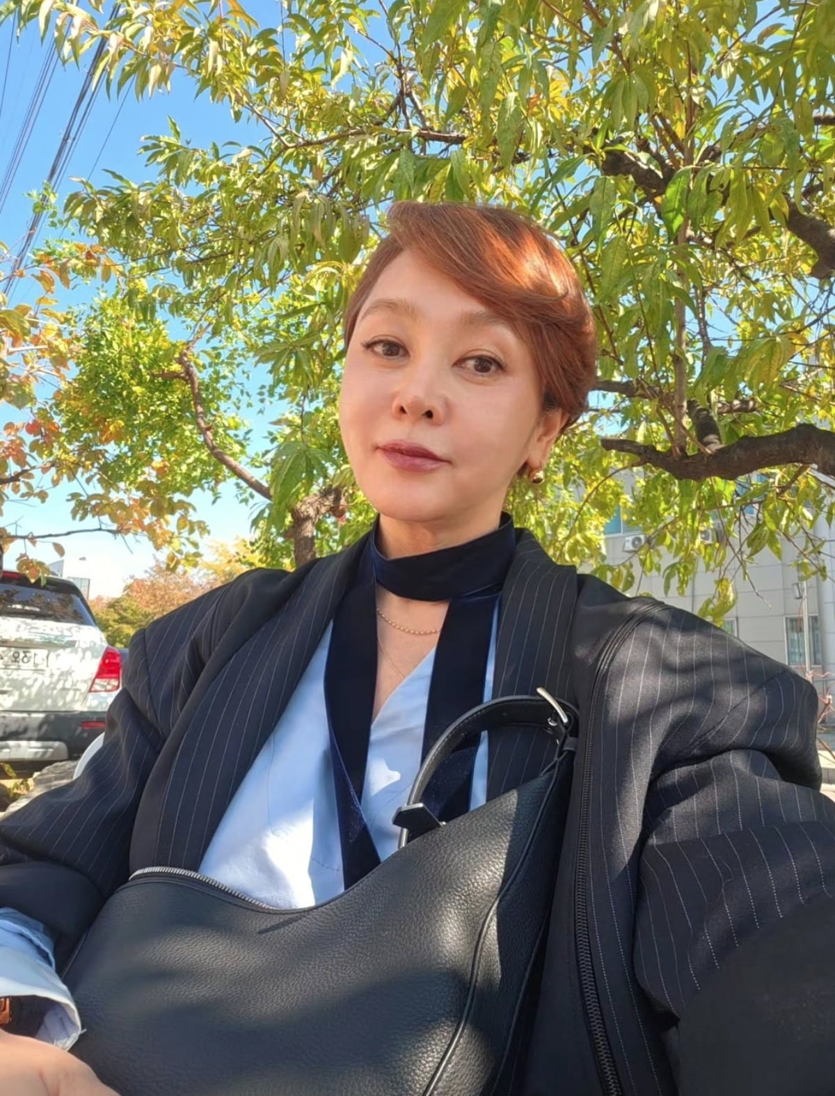木漏れ日の下で、淡いブルーの服とストライプのジャケット姿で自撮りするイ・スンヨン