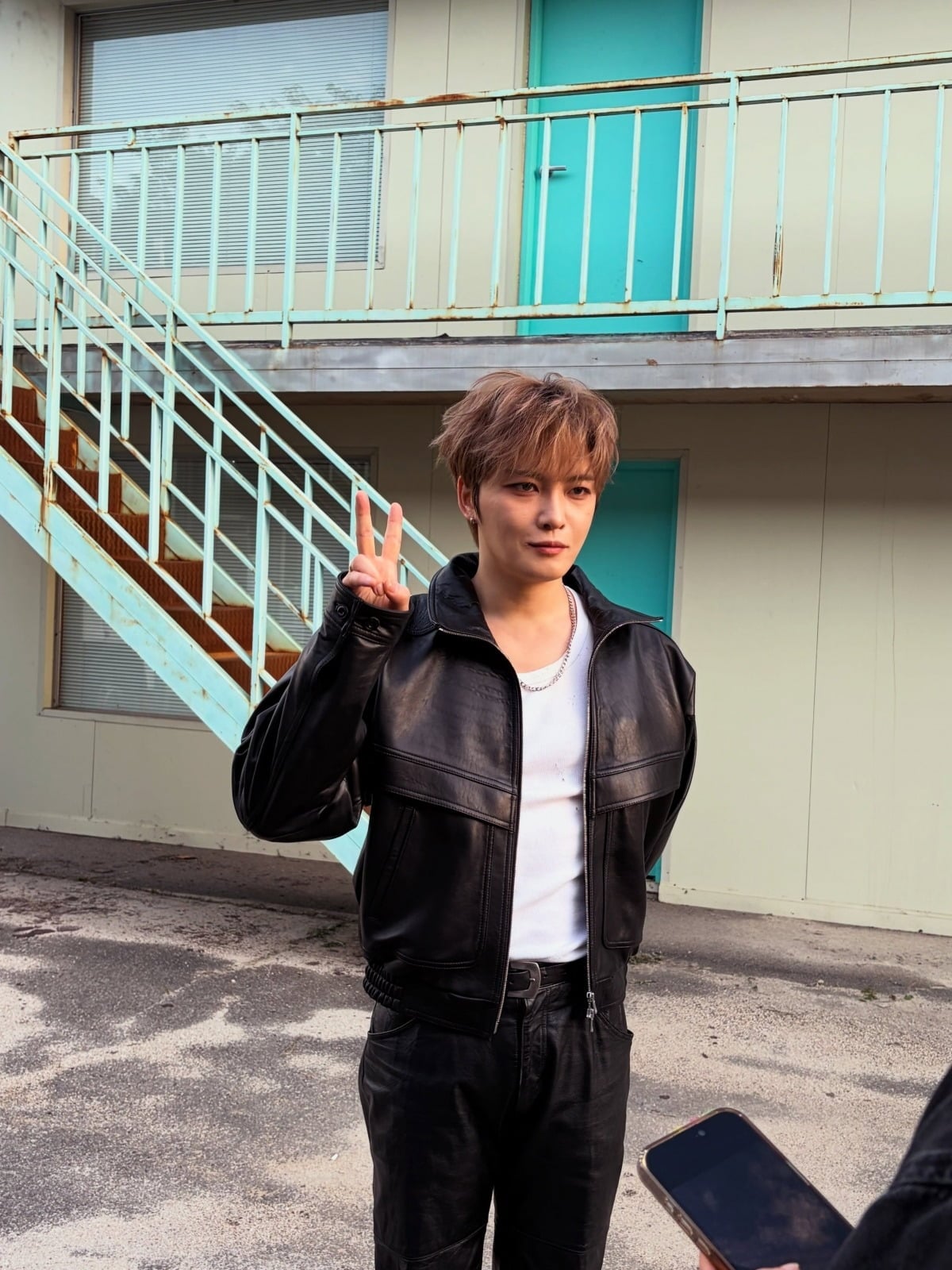 黒いレザージャケットとパンツに白いTシャツを合わせ、屋外でピースサインをするジェジュン