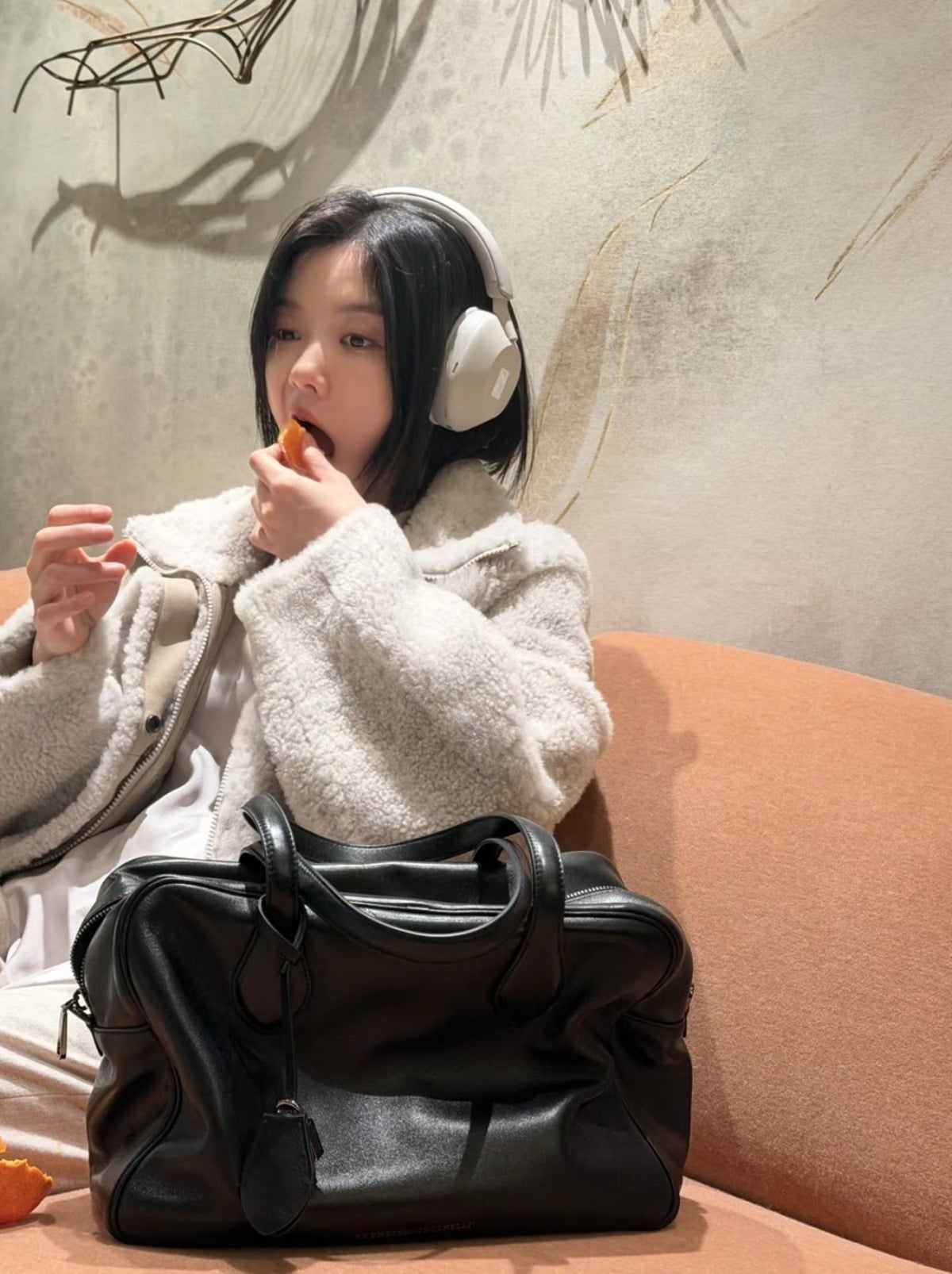 アイボリーのもこもこしたジャケットと白いヘッドホンを身につけ、みかんを食べるキム・ユジョン