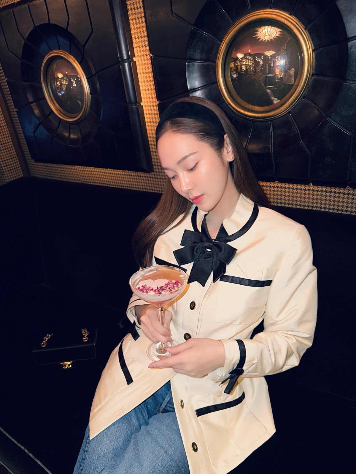 黒いリボン付きのアイボリージャケットを着て、ピンク色の飲み物を手にするジェシカ