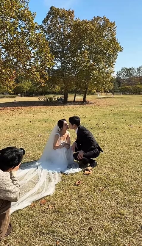 秋色の木々が広がる芝生で、ウェディングドレスと黒いスーツ姿でキスをする25期ヨンスとスンジャ。そばで撮影者がカメラを構えている