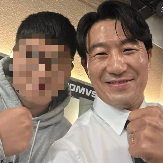 白いシャツと黒いネクタイ姿のサンチョルが、顔にぼかしの入った友人と並ぶ写真。