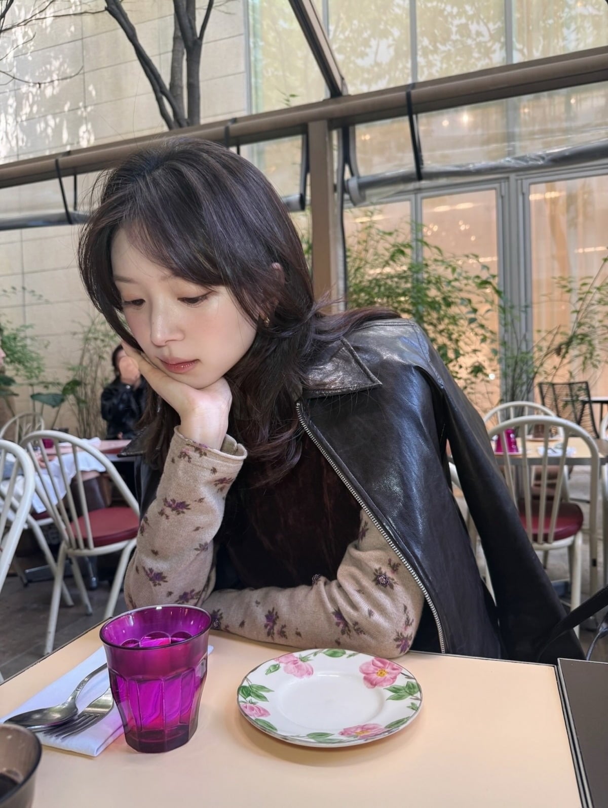 黒いレザージャケットを肩にかけ、カフェのテーブルで頬づえをつくキム・ジヨン