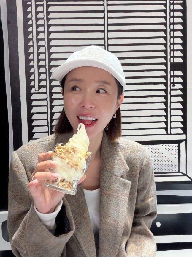白いキャップとブラウン系のジャケット姿で、アイスクリームを口元に近づけて笑うシンジ