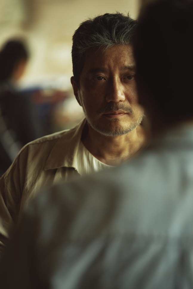 映画『悲しい熱帯』でサブを演じるキム・ミョンミン。薄暗い場所で正面を見つめる場面写真。