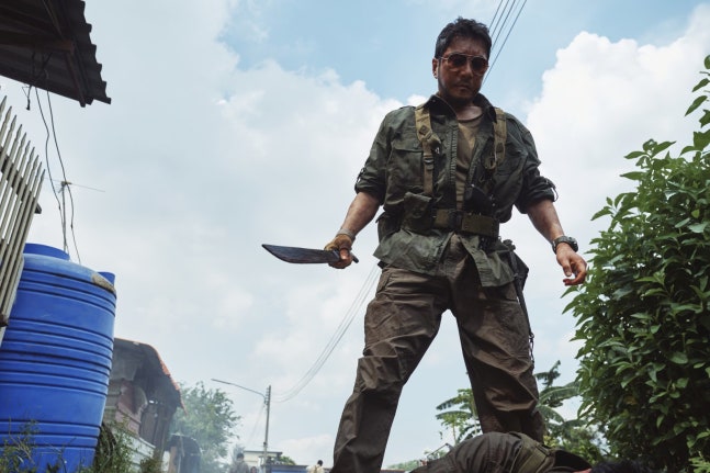 映画『悲しい熱帯』のキム・ミョンミン。野戦服姿でマチェーテを手にし、倒れた人物を見下ろす場面写真。