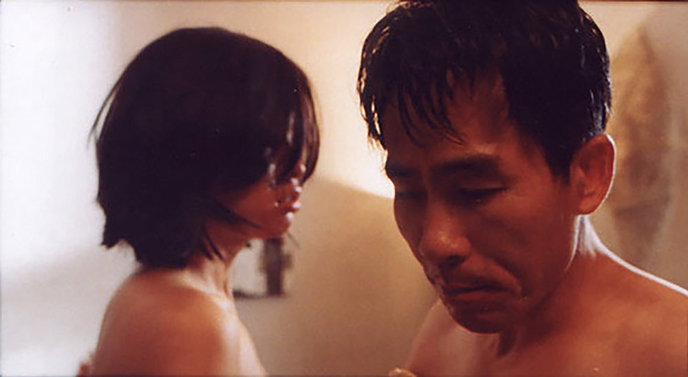 映画『嘘』のスチール写真。女性が男性のそばに座り、男性はうつむいている