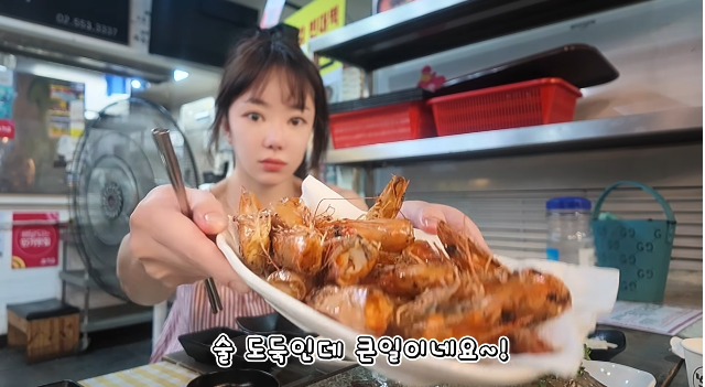飲食店でソ・ウが海鮮料理の皿をカメラに向けて見せる様子