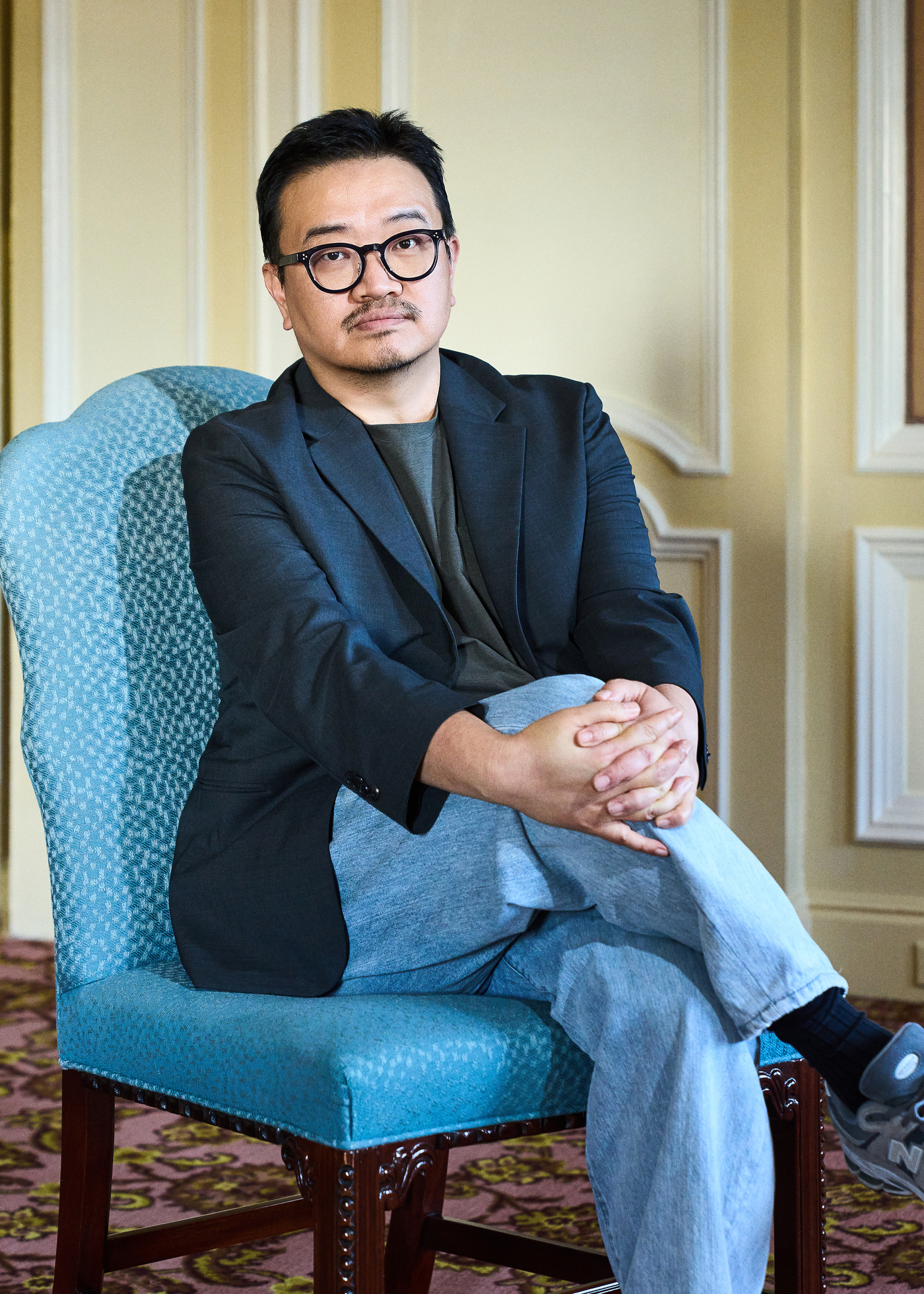 青い椅子に座り、黒縁眼鏡と紺色のジャケット姿で正面を見るヨン・サンホ監督