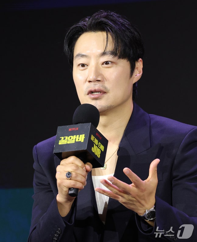 Netflixシリーズ『꿀알바』の制作発表会で、黒いスーツ姿のイ・ヒジュンが作品ロゴ入りのマイクを手に話す