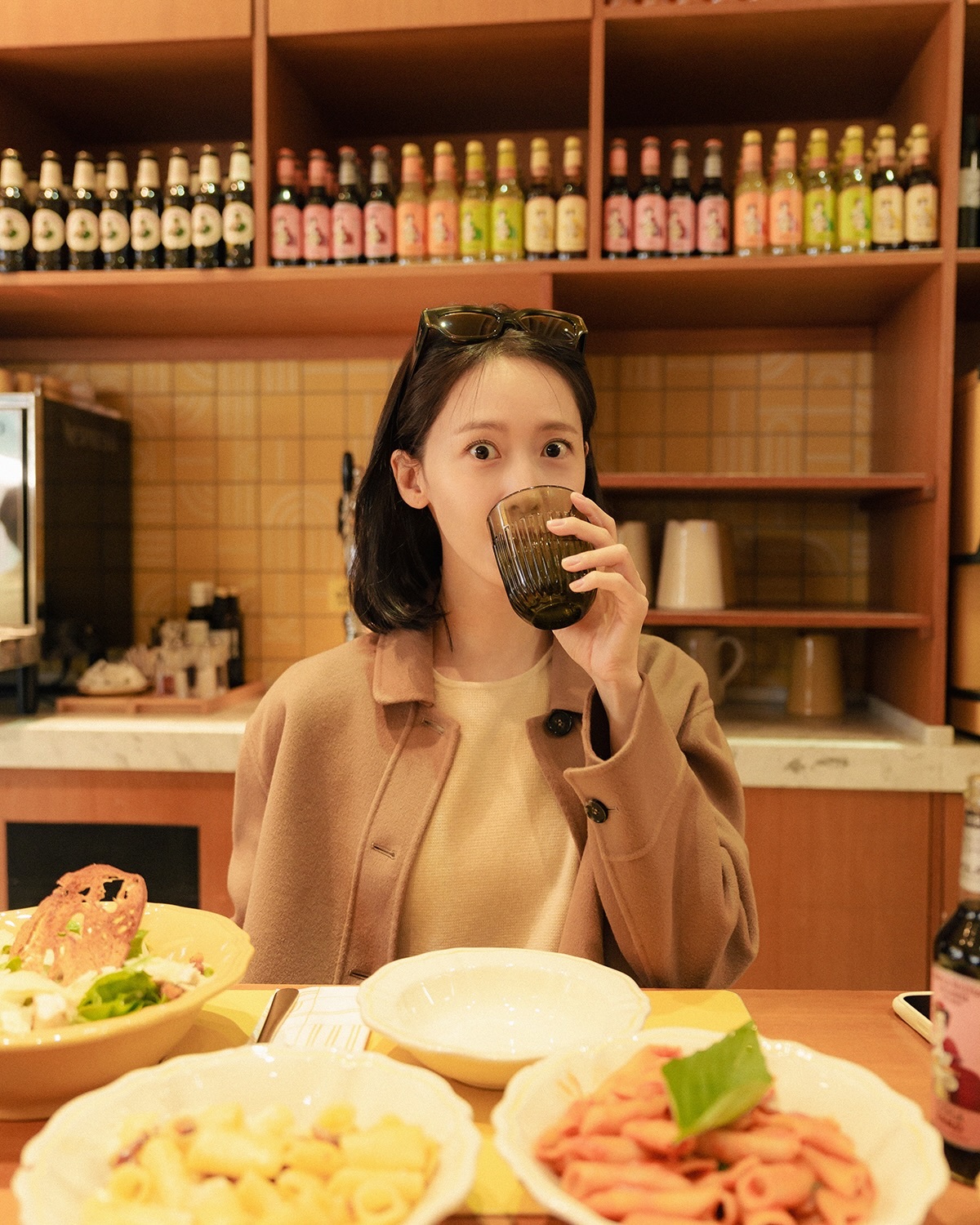 サングラスを頭に載せたユナが、茶色の上着姿でテーブル前に座り、目を大きく見開いてカメラを見る。