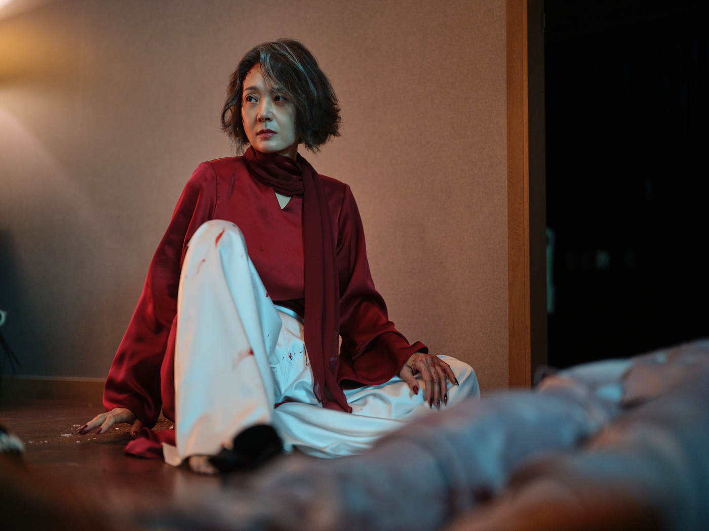 映画『完璧な家』のスチール。赤いトップスと白いパンツ姿のペ・ジョンオクが、薄暗い室内の床に座っている。